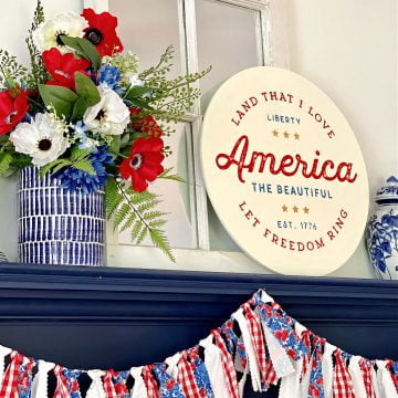 Round patriotic sign, floral display, and garland on a navy mantel
