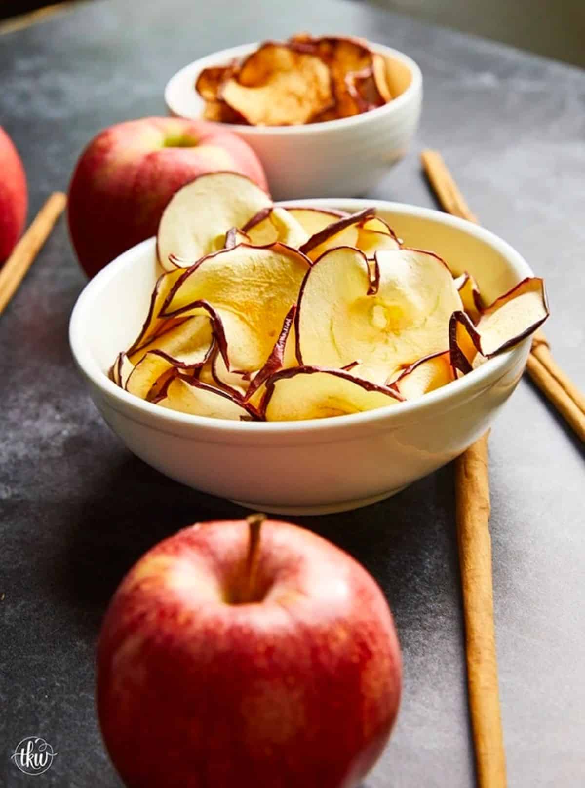 Homemade apple chips in a white bowl with whole apples on a table.