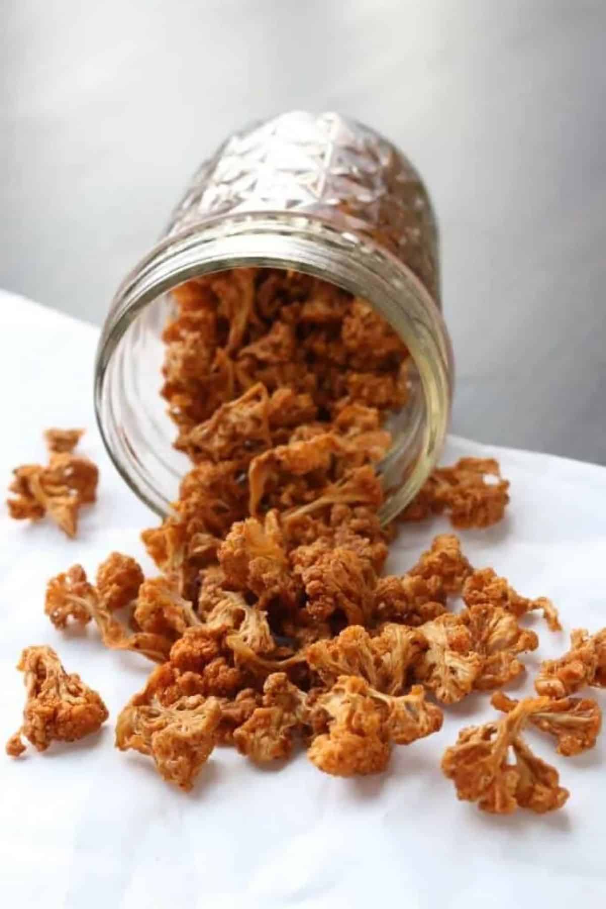 Dehydrated spiced cauliflower popcorn spilled from a glass jar.