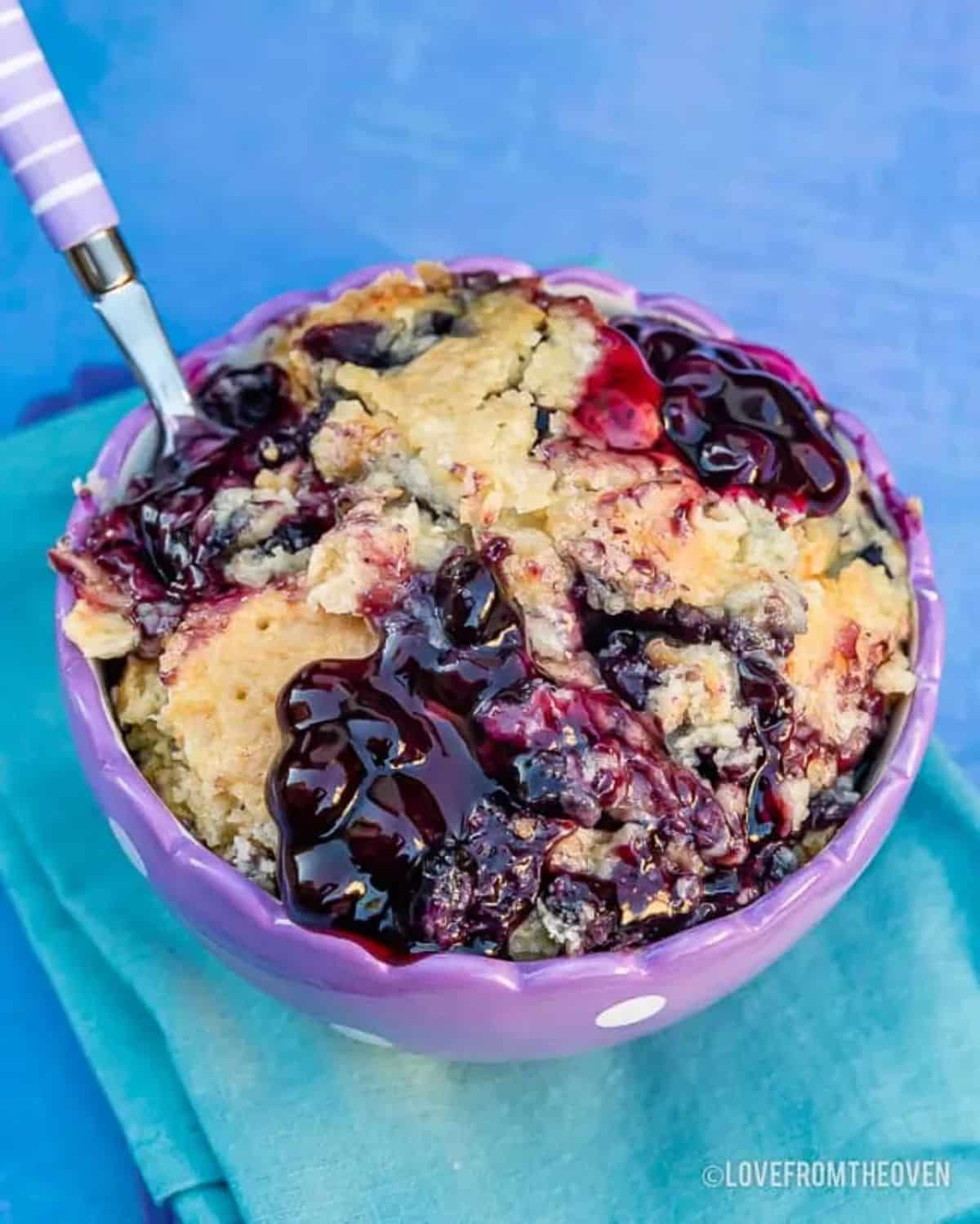 Blueberry dump cake in a purple bowl with a spoon.