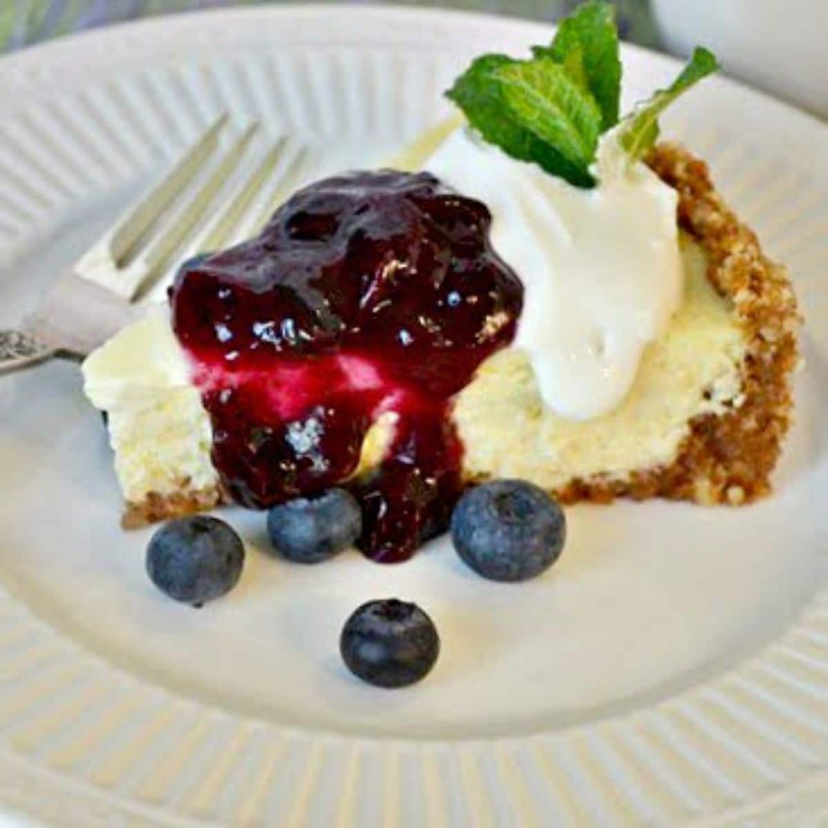 A piece of lemon cheesecake with blueberry topping on a white plate with a fork.