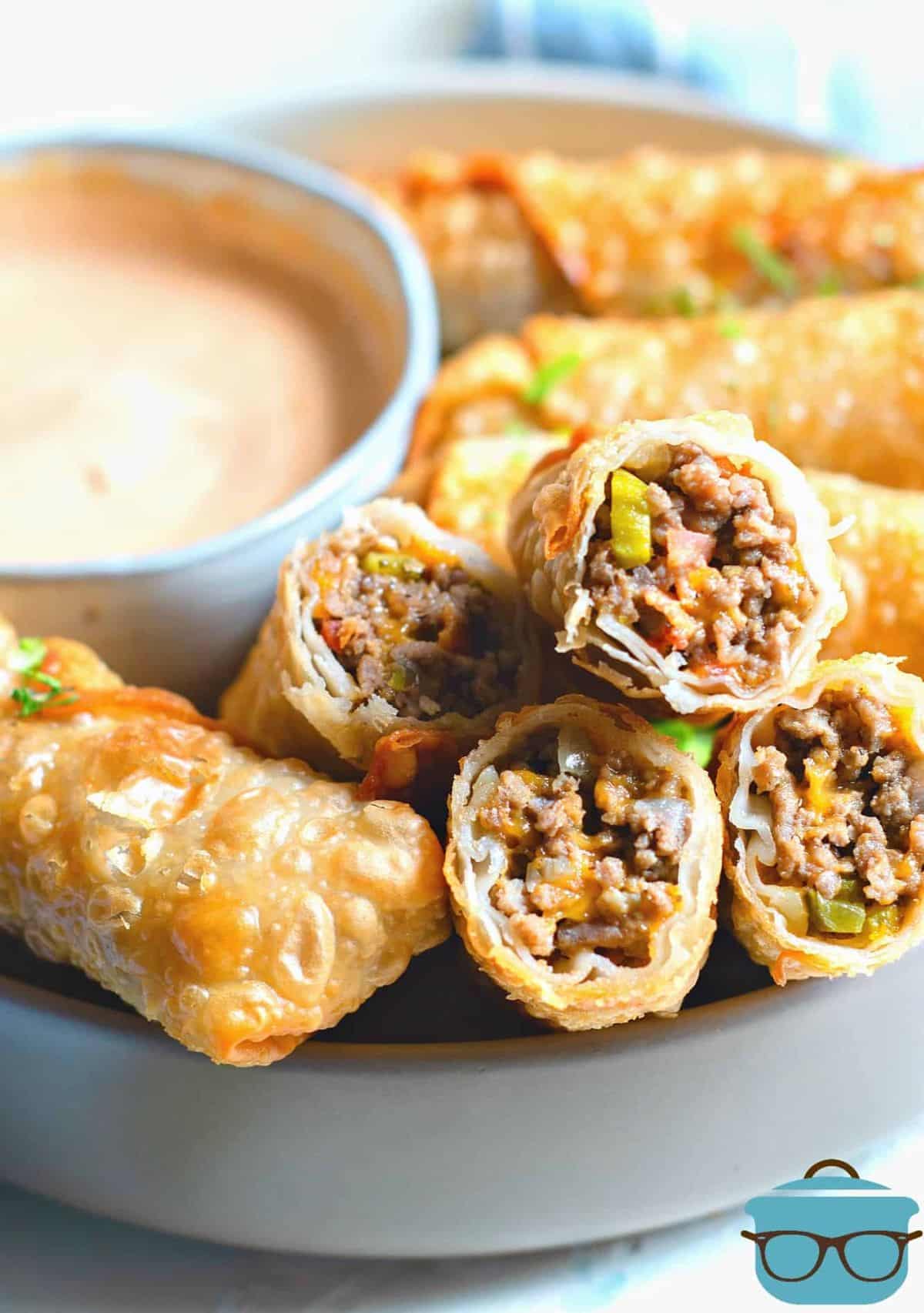 13. Cheeseburger egg rolls with a bowl of dip on a gray tray.