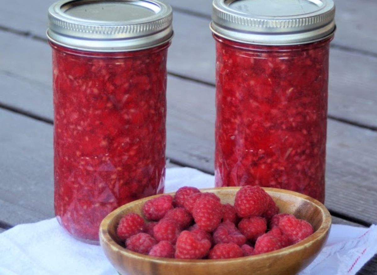 Raspberry pie filling in two glass jars with a bowl of ripe raspberries.