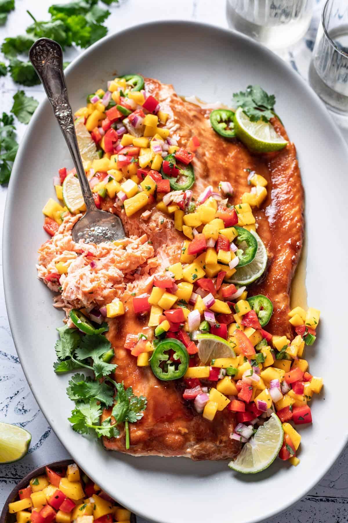 Baked salmon with mango salsa and chopped veggies on a white plate with a fork.
