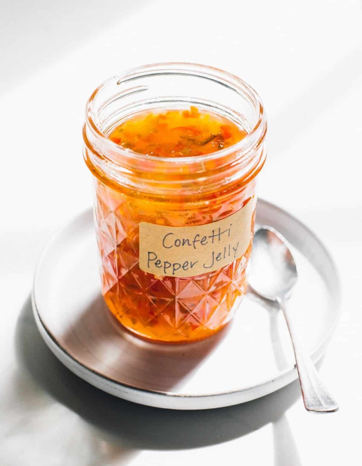 Homemade hot pepper jelly in a glass jar on a white plate with a spoon.