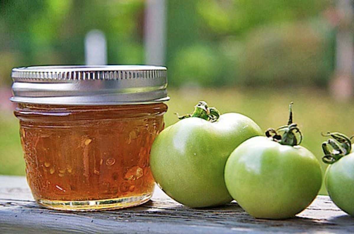 Green tomato marmalade canned in a glass jar with green tomatoes on a wooden board.