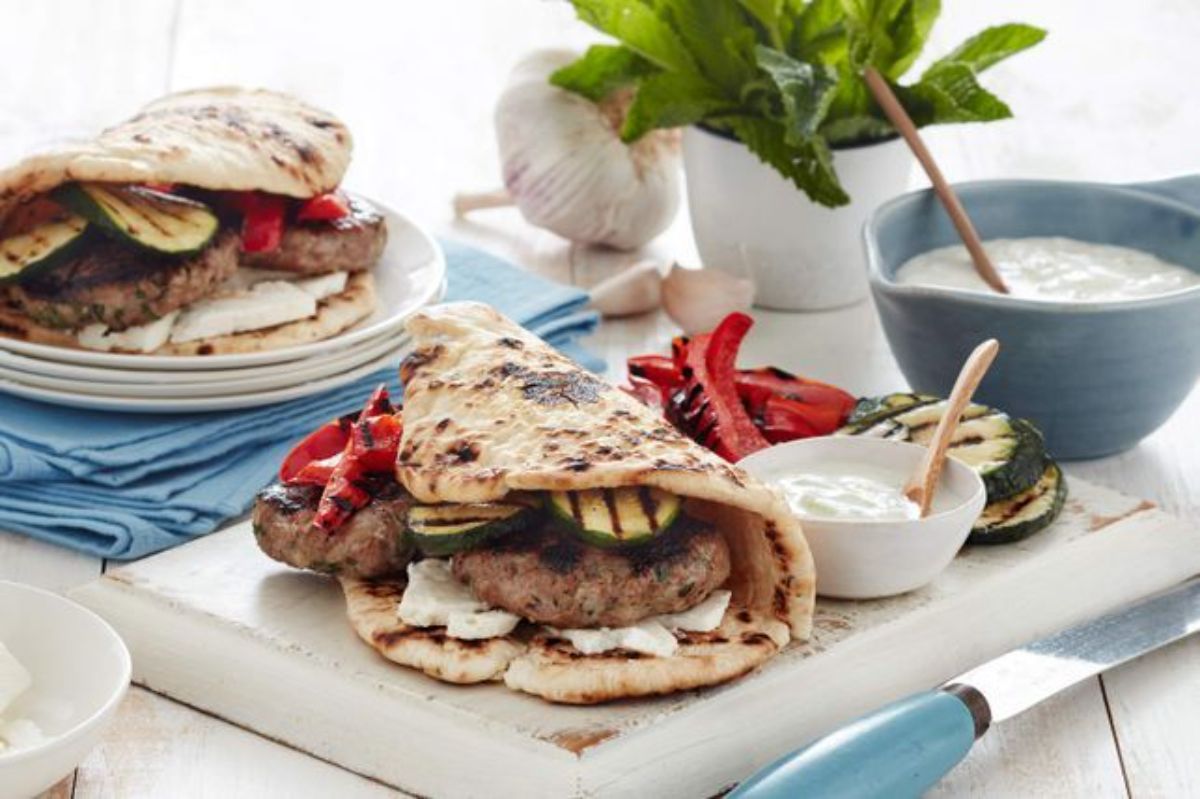 Barbecued lamb flatbread burgers with cucumber yogurt sauce on a white cutting board.