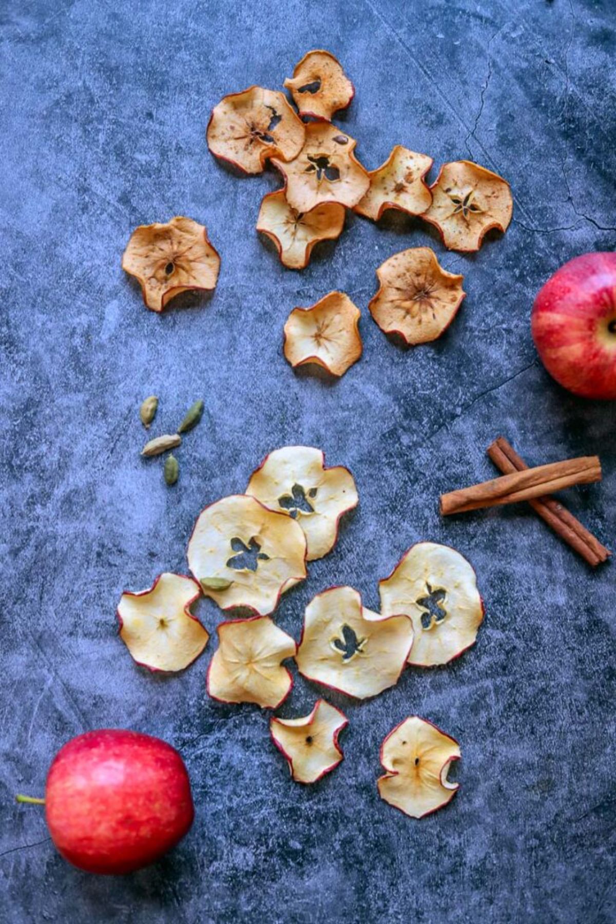Dehydrated apple chips scattered on a table with cinnamon sticks and two red apples.