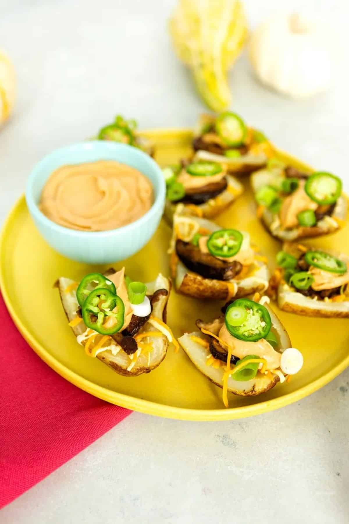 Vegan bacon loaded potato skins with a bowl of dip on a yellow tray.