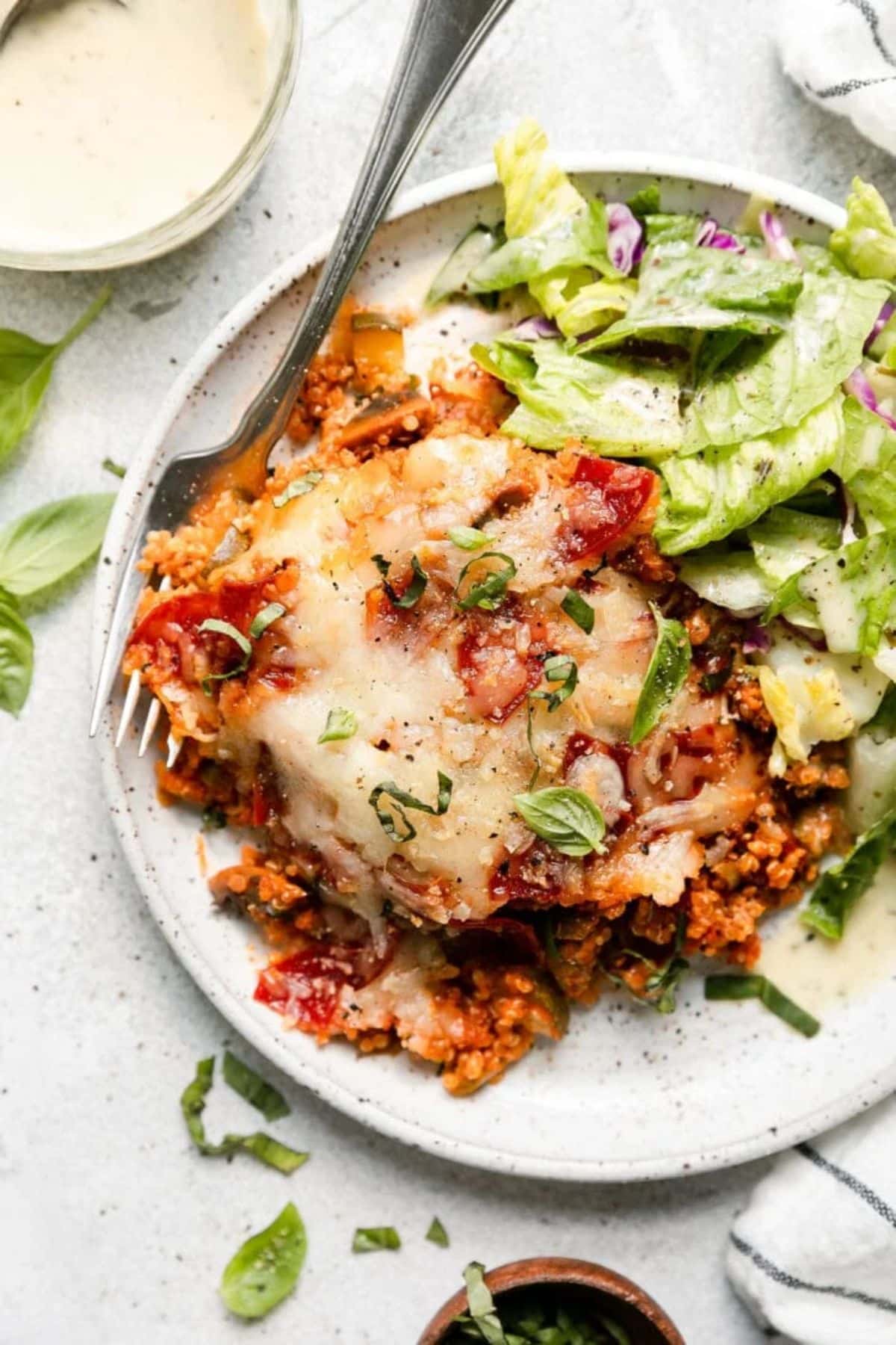 A piece of quinoa pizza on a plate with salad and a fork.