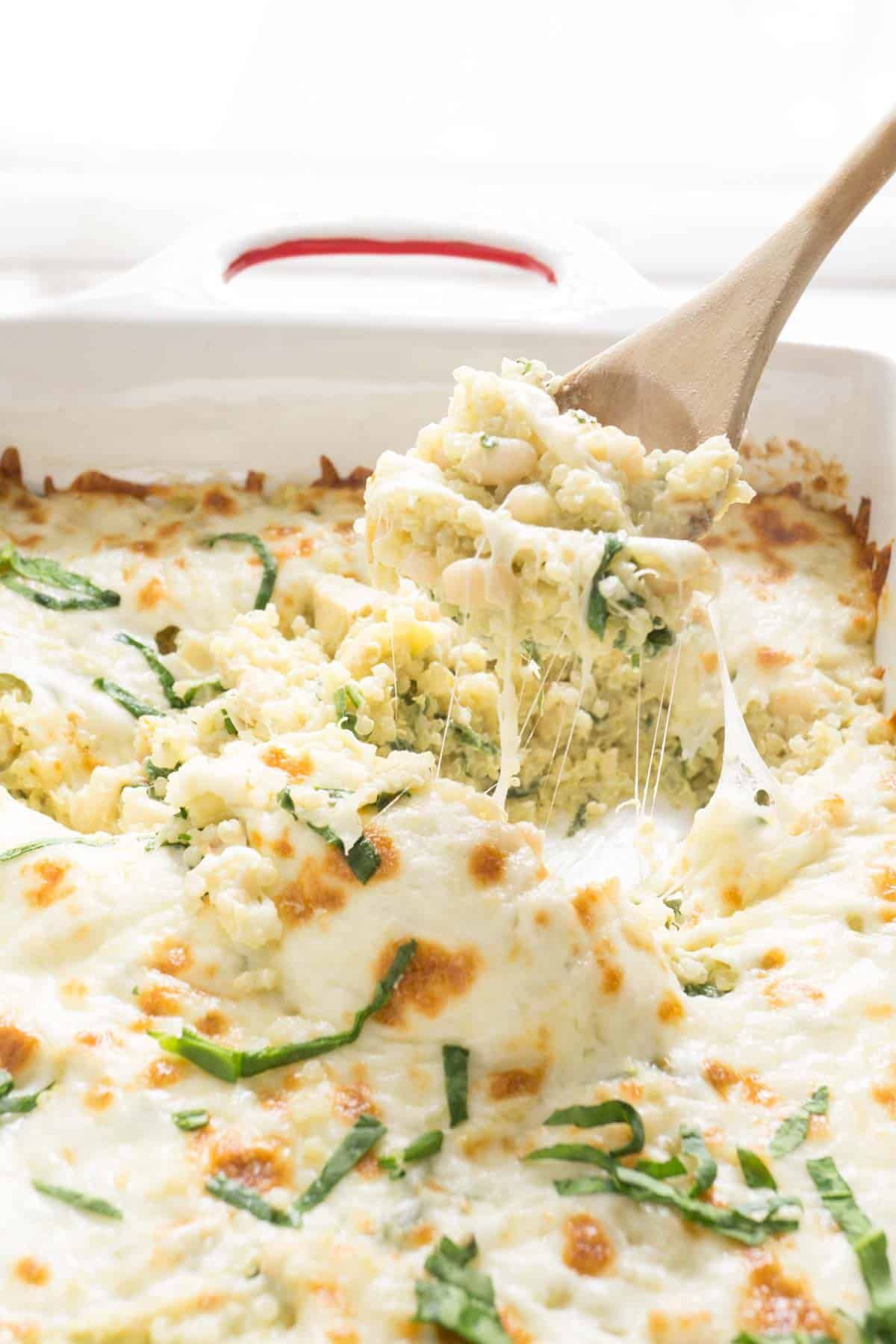 Juicy spinach and artichoke quinoa bake in a white casserole with a wooden spoon.