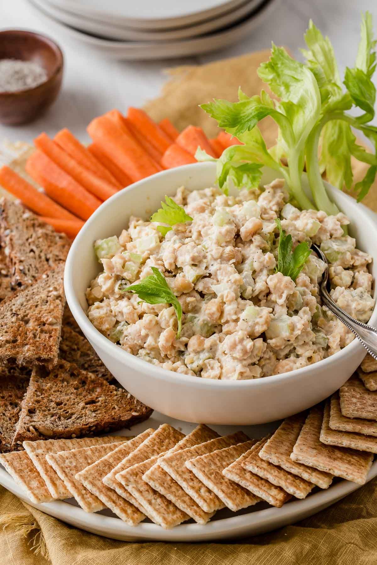 Tasty chickpea tuna salad in a white bowl with crackers and veggies on a tray.