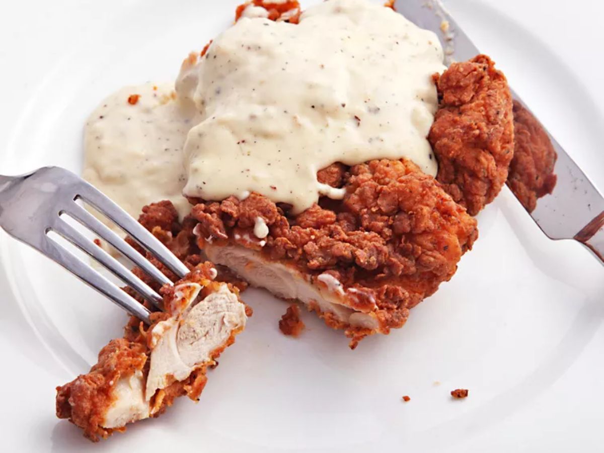 Crispy chicken-fried chicken with cream gravy on a white plate with cutlery.