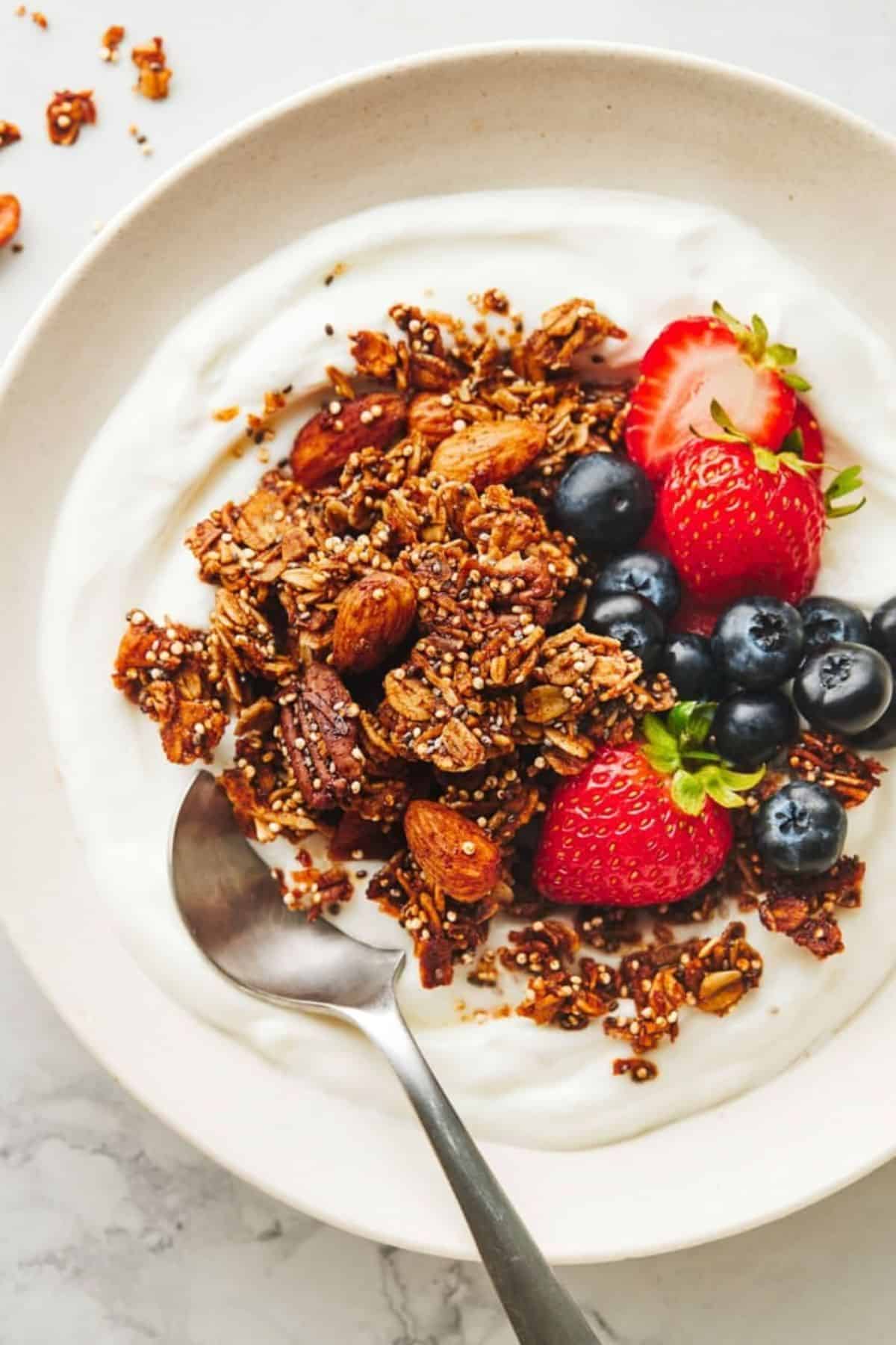 Crunchy maple quinoa granola with yogurt and fruits in a white bowl with a spoon.