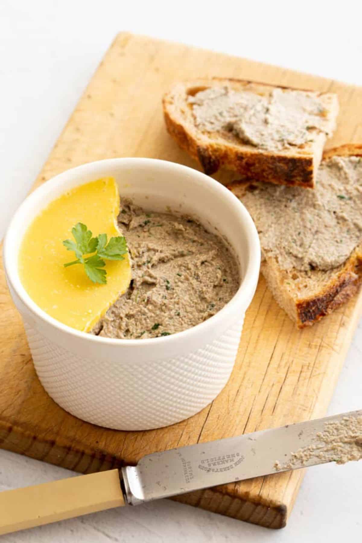 Creamy mushroom pate in a white bowl with pieces of bread and a knife on a wooden cutting board.