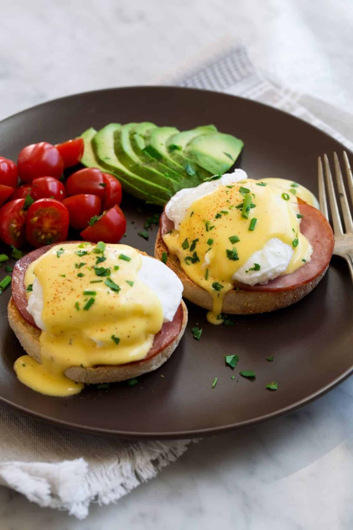 Mouth-watering eggs benedict with veggies on a black plate with a fork.