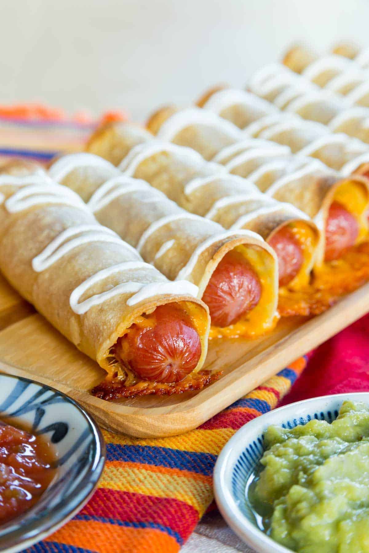 Crispy cheesy mexican hot dog taquitos on a wooden tray.