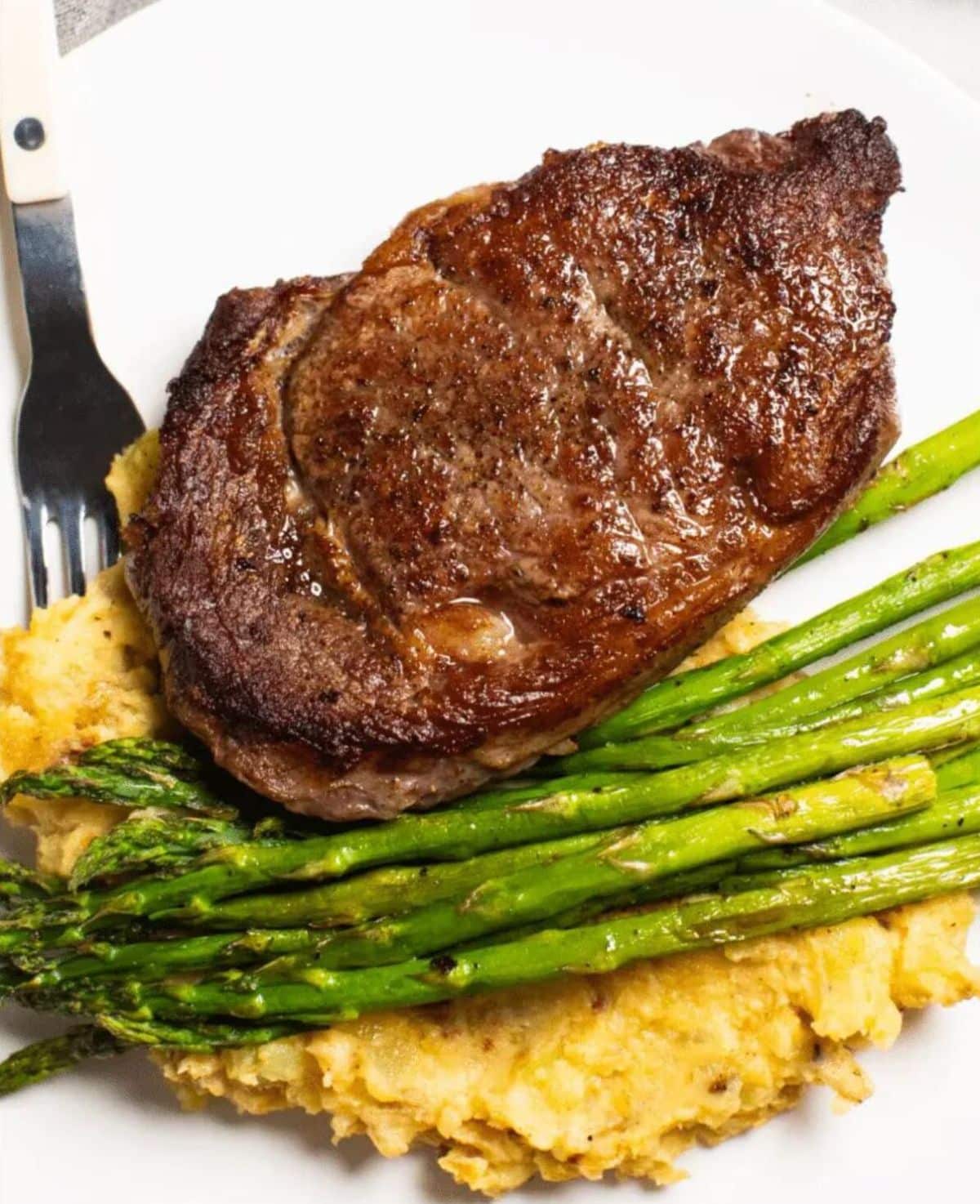 Simple bison ribeye dinner with mashed potatoes and asparagus on a white plate with a fork.