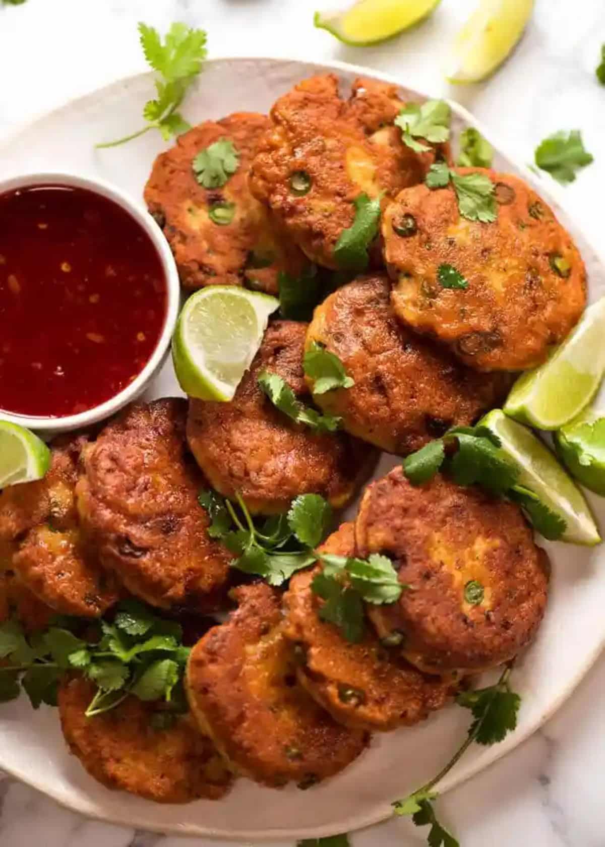 Scrumptious thai fish cakes with lime wedges and a bowl of dip on a white tray.