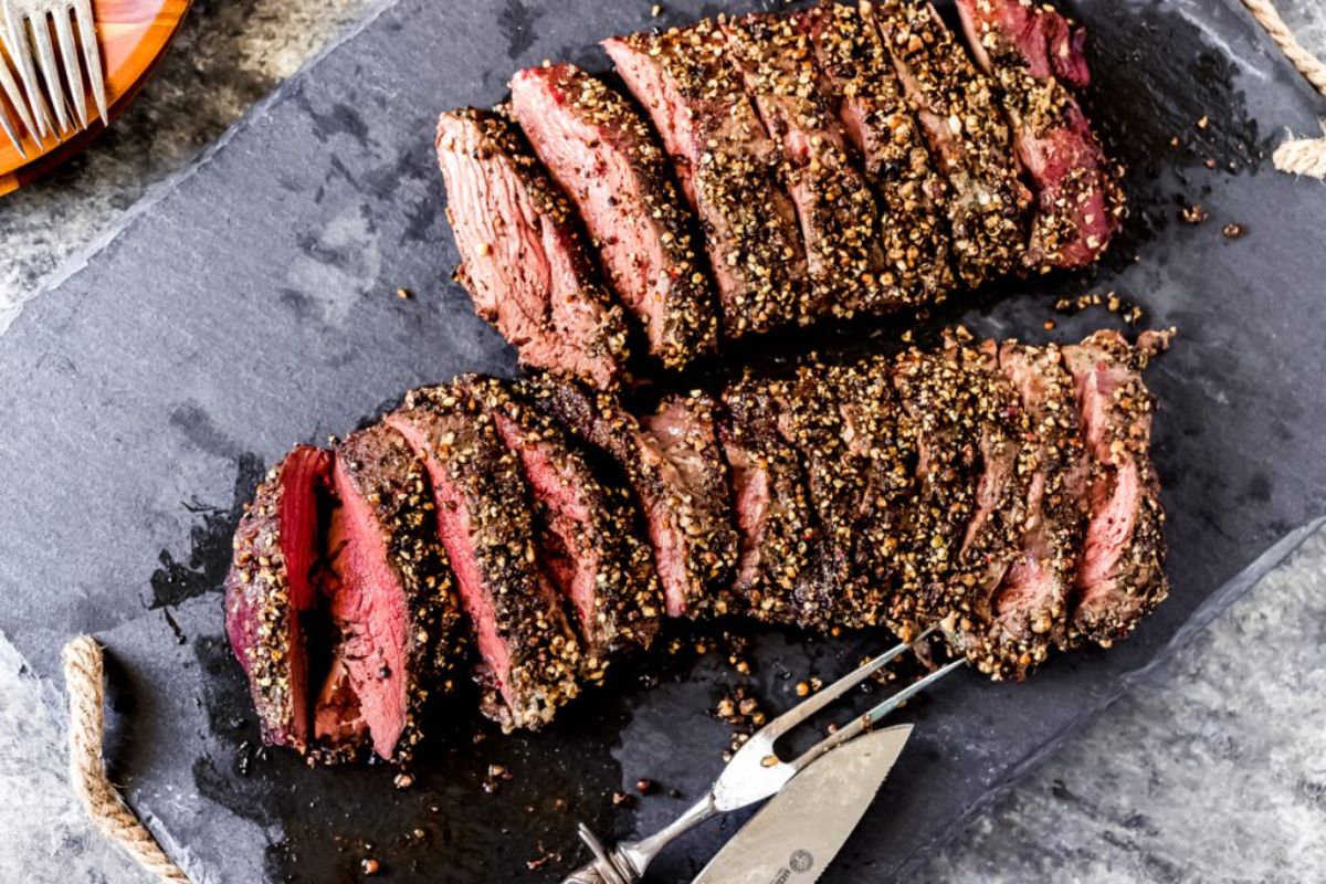 Mouth-watering peppercorn crusted elk tenderloin on a black tray with cutlery.