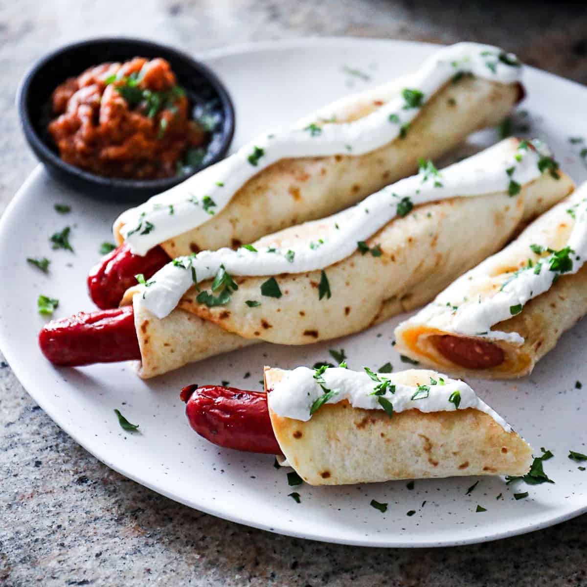 Tasteful hot dog taquitos on a white plate with a small bowl of dip.