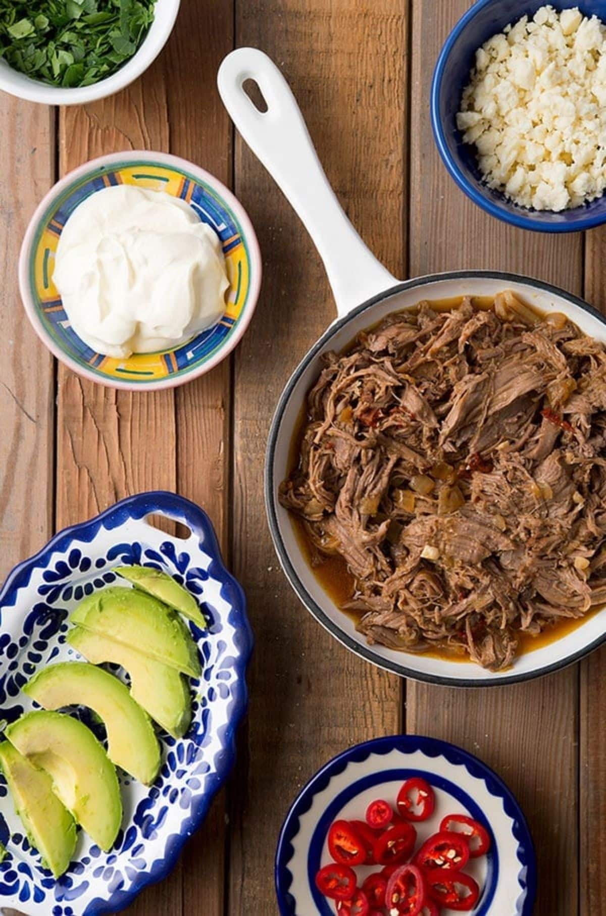 Juicy venison barbacoa in a skillet with ingredients on a wooden table.