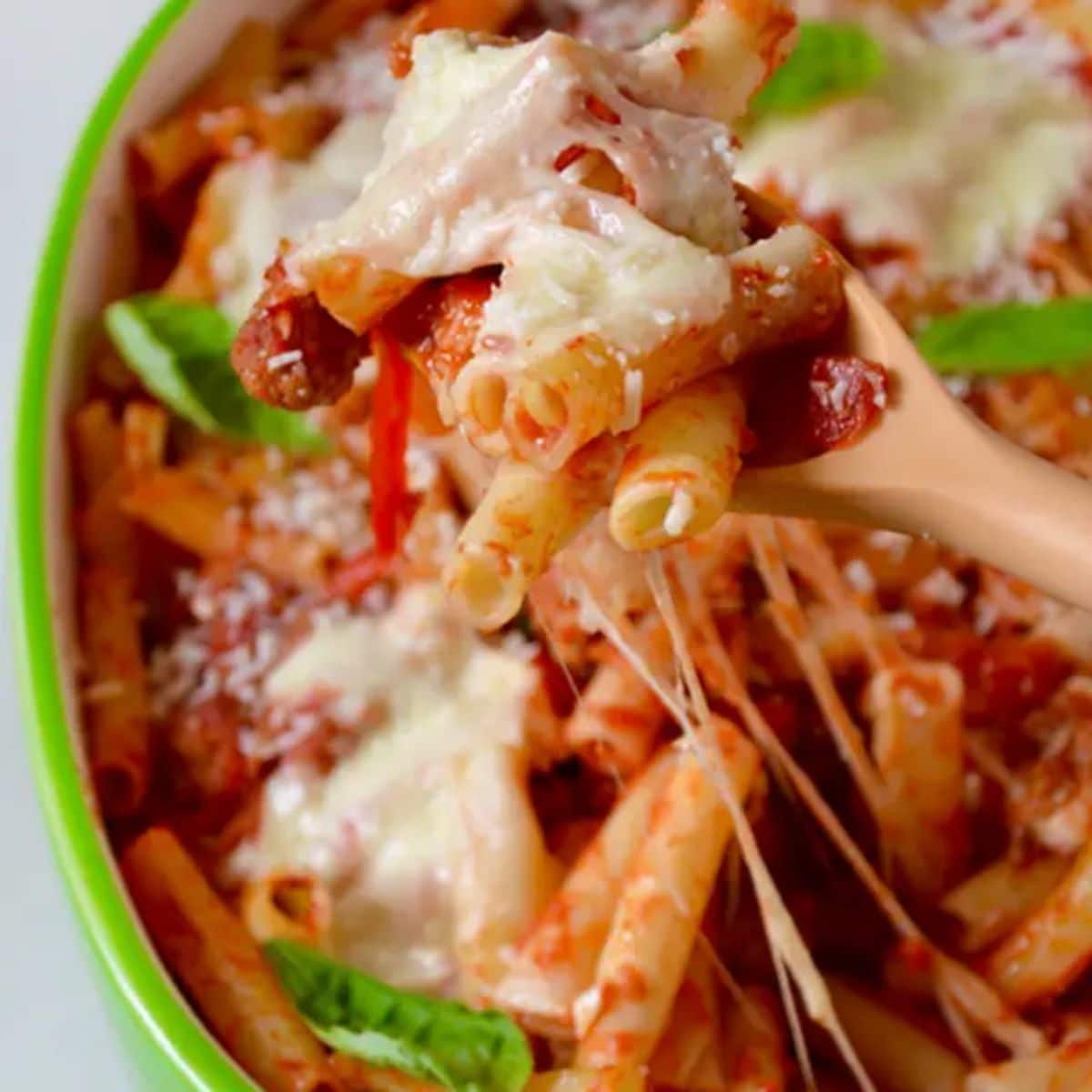 Juicy cheesy baked ziti with sausage and peppers in a casserole and on a wooden spoon.